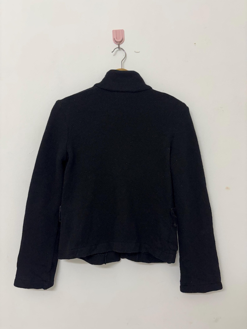 May include: A black jacket with a high collar, hanging on a hook. The jacket has long sleeves and a simple, classic design. The fabric appears to be a medium-weight material, suitable for cooler weather.