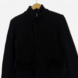 May include: A black jacket with a high collar and a front zipper. The jacket has two square pockets with a dark, textured material. The jacket is hanging on a wooden hanger against a white background.