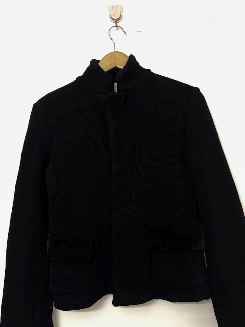 May include: A black jacket with a high collar and a front zipper. The jacket has two square pockets with a dark, textured material. The jacket is hanging on a wooden hanger against a white background.
