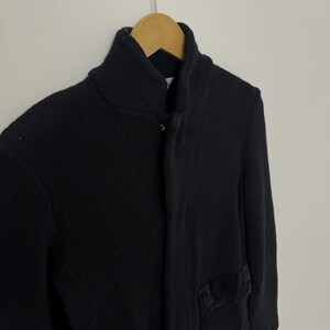 May include: A black knit cardigan with a high collar and two front pockets. The cardigan is hanging on a wooden hanger against a white background. The pockets have a dark, textured trim.