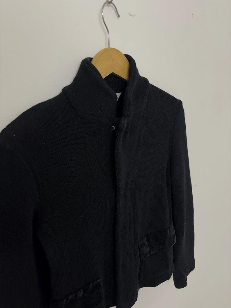 May include: A black knit cardigan with a high collar and two front pockets. The cardigan is hanging on a wooden hanger against a white background. The pockets have a dark, textured trim.