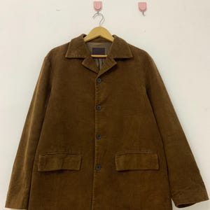 May include: A brown corduroy blazer with a button-up front and two pockets. The blazer is hanging on a wooden hanger.