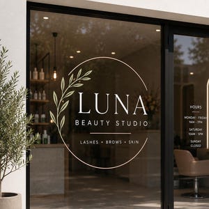 May include: A glass storefront window featuring a white circular logo with a leaf branch design and the text LUNA BEAUTY STUDIO LASHES BROWS SKIN. To the right, white text displays hours: MONDAY-FRIDAY 9AM-7PM, SATURDAY 10AM-5PM, SUNDAY CLOSED.