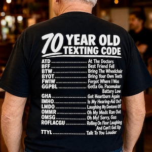 May include: A black t-shirt featuring white text that reads 70 YEAR OLD TEXTING CODE followed by a list of abbreviations and their meanings, including ATD At The Doctors, BFF Best Friend Fell, BTW Bring The Wheelchair, and other humorous phrases.