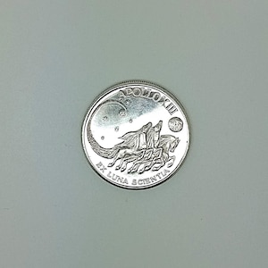 Può includere: Una moneta d'argento con un design a rilievo dettagliato e un bordo zigrinato. La moneta presenta la scritta "APOLLO XIII" e "EX LUNA SCIENTIA". L'immagine raffigura tre cavalli con criniere fluenti, una luna crescente, stelle e una luna piena.