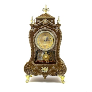 May include: An ornate gold-toned clock with a quartz movement. The clock has a decorative, carved wooden case with a glass front. The clock face is white with black Roman numeral hour markers and black hands.