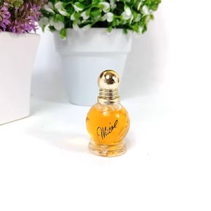 Può includere: Piccola bottiglia di profumo rotonda con tappo dorato e base trasparente. La bottiglia contiene un liquido dorato e presenta la scritta "Micol" in corsivo. Su sfondo bianco, con piante in vaso sullo sfondo.
