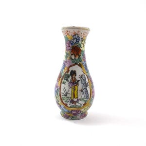 May include: A small, white porcelain vase with a pear-shaped body and a narrow neck. The vase is decorated with a colourful floral pattern and a scene of a woman standing in a garden. The vase has a gold trim around the rim and base.