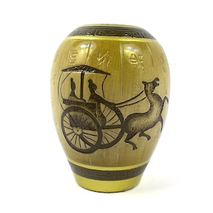May include: A gold-coloured ceramic vase featuring a dark brown illustration of a horse-drawn carriage. The vase has a cylindrical shape with decorative bands at the top and bottom. The background is white.
