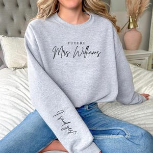 May include: A pale grey sweatshirt featuring the script text "FUTURE Mrs. Williams" across the chest. The sleeve displays the text "I said yes". The sweatshirt is paired with blue jeans.