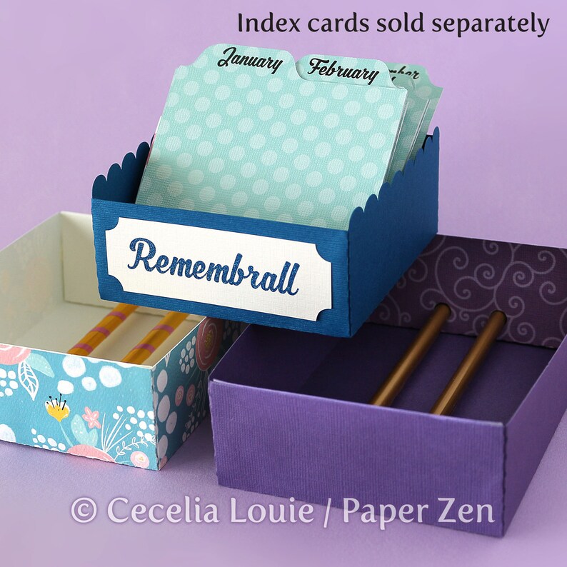 Puede incluir: Una caja de papel azul y blanca con un borde festoneado y una etiqueta que dice "Rememberall". La caja est&aacute; llena de tarjetas de &iacute;ndice etiquetadas "Enero", "Febrero" y "Marzo". La caja est&aacute; encima de una caja morada con dos l&aacute;pices dentro.