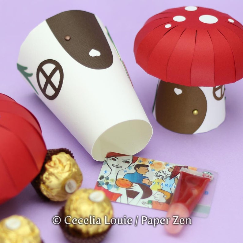 May include: A red and white paper mushroom with a brown base and a white paper house with a brown roof and a window. There are three gold chocolate balls and a red lip gloss tube on a purple surface.