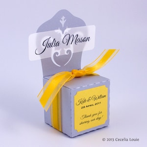 May include: A gray and yellow wedding favor box with a white tag that says "Julia Mason". The box has a yellow ribbon and a yellow label that says "Kate & William 29 APRIL 2011 Thank you for sharing our day!"