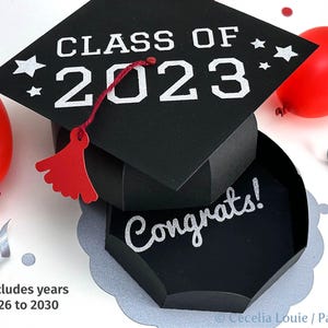 May include: A black graduation cap-shaped gift box with the text "CLASS OF 2023" and "Congrats!". The box is decorated with red and silver accents, including balloons and confetti. Includes years 2026 to 2030.