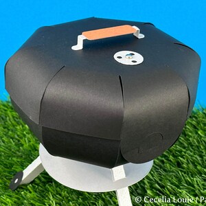 Puede incluir: Un modelo de papel de una peque&ntilde;a parrilla de carb&oacute;n negro con un asa marr&oacute;n en la tapa. La parrilla est&aacute; sobre una superficie de hierba verde.
