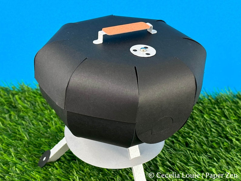 Puede incluir: Un modelo de papel de una peque&ntilde;a parrilla de carb&oacute;n negro con un asa marr&oacute;n en la tapa. La parrilla est&aacute; sobre una superficie de hierba verde.