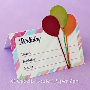 Könnte beinhalten: Eine gefaltete Geburtstagskarte mit einem farbenfrohen diagonalen Streifenmuster. Die Karte zeigt das Wort "Birthday" in einer Schreibschrift, mit Linien für Name, Geburtstag und Notizen. Drei Papierballons in Rot, Grün und Orange sind mit Schnur befestigt.