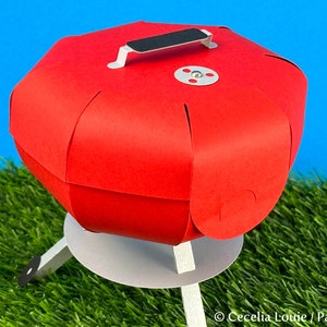 Puede incluir: Un modelo de papel de una peque&ntilde;a barbacoa redonda de color rojo con un asa negra y una base blanca. La barbacoa est&aacute; sobre una superficie de hierba verde.