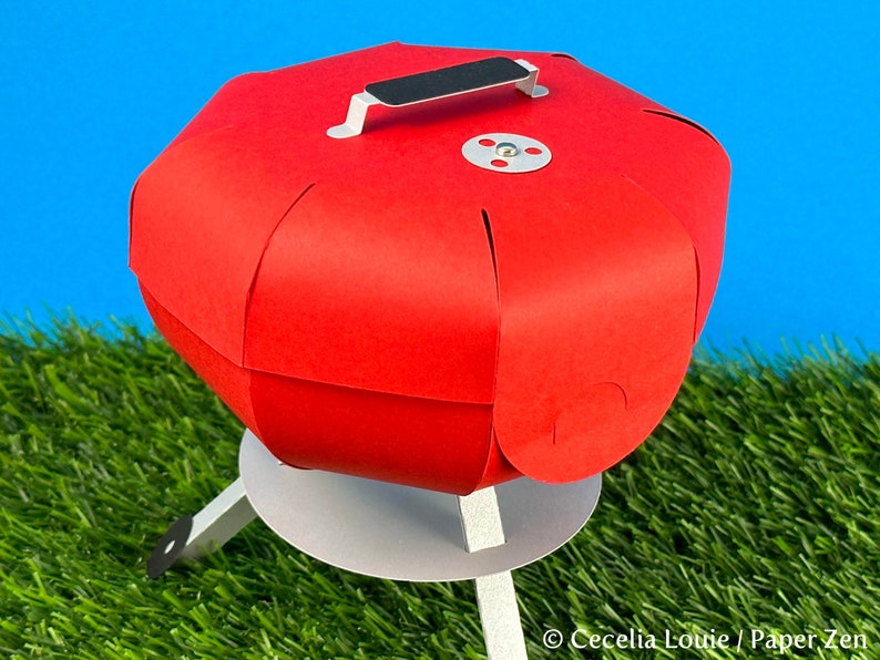 Puede incluir: Un modelo de papel de una peque&ntilde;a barbacoa redonda de color rojo con un asa negra y una base blanca. La barbacoa est&aacute; sobre una superficie de hierba verde.