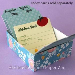 Puede incluir: Una caja de color azul y blanco con estampado floral y un dise&ntilde;o floral amarillo, rosa y blanco. La caja est&aacute; abierta y tiene una tarjeta amarilla en el interior con el texto "Heirloom Seeds" y las palabras "Variety", "When to Sow", "Depth" y "Rows".