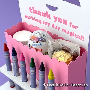 Puede incluir: Caja de cart&oacute;n rosa con borde festoneado, llena de l&aacute;pices de colores, un huevo de chocolate y un peque&ntilde;o recipiente blanco. La caja tiene una etiqueta blanca adjunta que dice "thank you for making my day magical!"