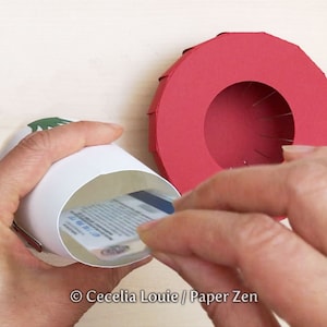 May include: A white paper cylinder with a green tree design on the top is being used to hold a gift card. The cylinder is being placed inside a red paper ring with a hole in the center.