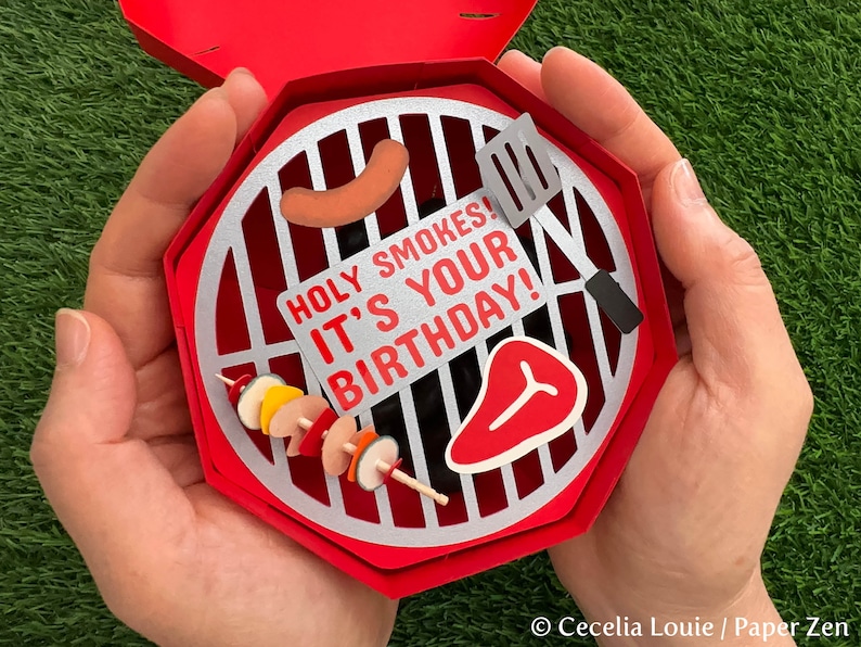 Puede incluir: Una tarjeta de cumplea&ntilde;os en forma de parrilla roja y plateada con el texto "Holy Smokes! It's Your Birthday!" en el interior.