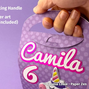 Puede incluir: Caja de regalo de papel morado con un patr&oacute;n de lunares blancos. La caja tiene un asa de cierre y una etiqueta de nombre "Camila" rosa y blanca con un n&uacute;mero 6 blanco. La caja tambi&eacute;n presenta una ilustraci&oacute;n de unicornio con el texto "Cecelia Louie. Paper Zen".
