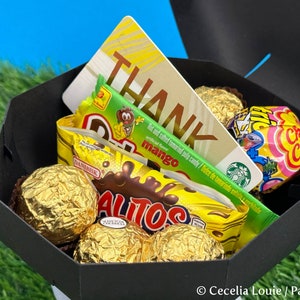 Puede incluir: Una caja de regalo negra llena de varios dulces, incluyendo caramelos cubiertos de chocolate, una barra de caramelo verde y amarilla, un caramelo rosa y amarillo y una tarjeta de regalo con la palabra "THANK" en ella.