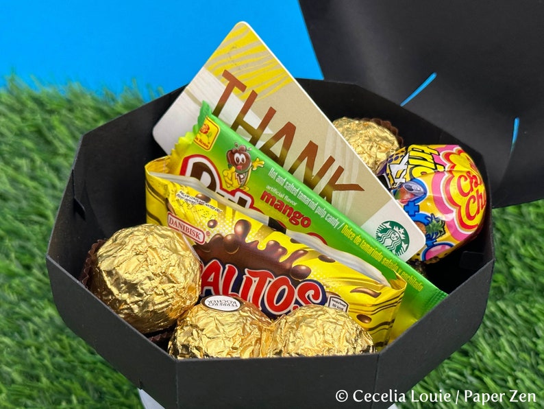 Puede incluir: Una caja de regalo negra llena de varios dulces, incluyendo caramelos cubiertos de chocolate, una barra de caramelo verde y amarilla, un caramelo rosa y amarillo y una tarjeta de regalo con la palabra "THANK" en ella.