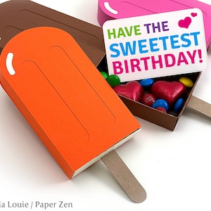 May include: Three popsicle shaped boxes, one orange, one brown, and one pink. The pink box is open and has a white card inside that says "Have the Sweetest Birthday!" with a red heart. The boxes are filled with colorful candies.