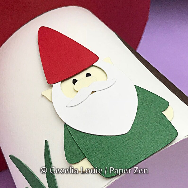 May include: A papercraft gnome with a red hat, white beard, and green shirt. The gnome is partially visible against a white background with green grass.