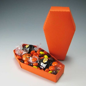 Puede incluir: Caja de cartón naranja en forma de ataúd llena de dulces de Halloween surtidos, incluyendo dulces de chocolate y mantequilla de cacahuete.