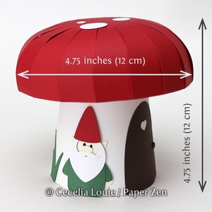 May include: A red and white paper mushroom house with a white door and a heart-shaped window. A green and red gnome figure is attached to the side of the house. The mushroom is 4.75 inches (12 cm) wide and 4.75 inches (12 cm) tall.