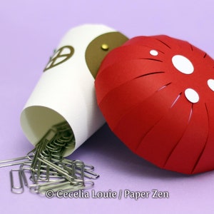 May include: A red and white paper mushroom with a white paper cup underneath. The cup is filled with silver paper clips. The mushroom and cup are on a purple surface.