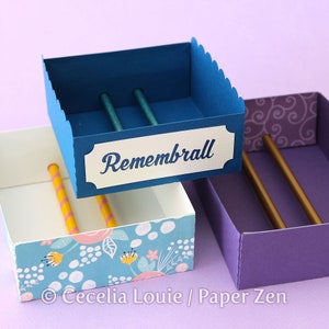 Puede incluir: Tres cajas apiladas con diferentes patrones y colores. La caja superior es azul con una etiqueta blanca que dice "Rememberall". La caja del medio es azul y blanca con un patr&oacute;n floral. La caja inferior es morada con un patr&oacute;n de espiral.