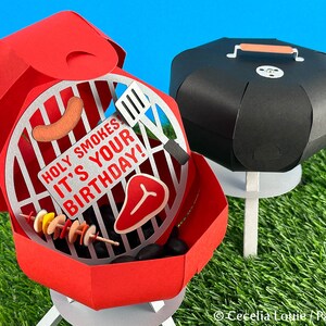 Puede incluir: Barbacoa de papel roja y negra con una tapa que se abre para revelar una parrilla con una salchicha, un bistec y brochetas de comida. La barbacoa tiene un letrero que dice "Holy Smokes It's Your Birthday!"