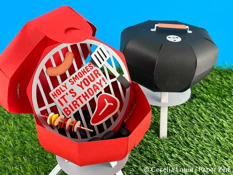 Puede incluir: Barbacoa de papel roja y negra con una tapa que se abre para revelar una parrilla con una salchicha, un bistec y brochetas de comida. La barbacoa tiene un letrero que dice "Holy Smokes It's Your Birthday!"