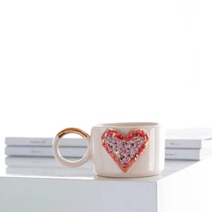 Könnte beinhalten: Cremefarbene Keramiktasse mit goldenem Henkel und herzförmigem Design. Das Herz ist mit rosa und lila strukturierten Elementen und goldenen Akzenten gefüllt. Die Tasse steht auf einer weißen Oberfläche.