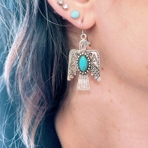 May include: Silver tone earrings with a turquoise stone in the shape of a stylized bird. The earrings are dangling and have a detailed design.