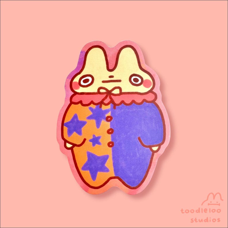 May include: A whimsical sticker featuring a cartoon rabbit character in a colorful outfit. The rabbit wears a red ruffled collar and a two-toned outfit, orange with blue stars on one side and solid blue on the other. The sticker has the text "toodleloo studios".