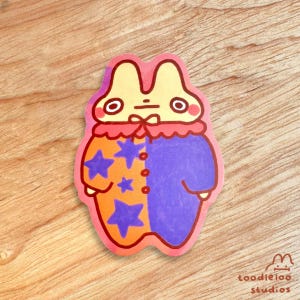 May include: A sticker featuring a cartoon rabbit-like character in a colorful outfit. The character has a yellow head, red collar, and a two-toned body, orange with purple stars on one side and purple on the other. The sticker has a pink border and the text "toodleloo studios".