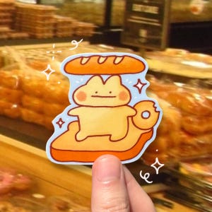 May include: A sticker featuring a cartoon character made of bread. The character is yellow with a smiling face, and is sandwiched between two pieces of bread. The sticker has a blue background and is surrounded by decorative sparkles.