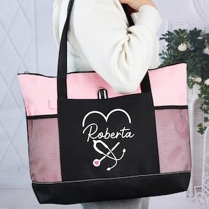 May include: A black and pink tote bag with the name "Roberta" and a stethoscope design. The bag has a black base, pink top and sides, and mesh side pockets. The bag is held by a person.