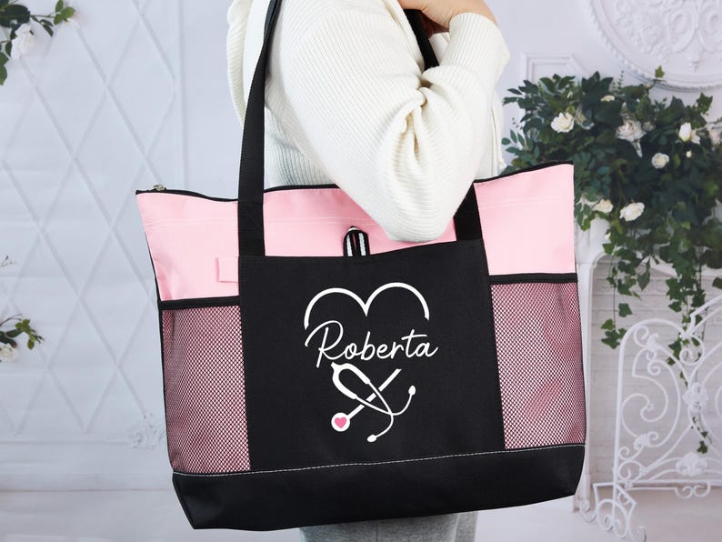 May include: A black and pink tote bag with the name "Roberta" and a stethoscope design. The bag has a black base, pink top and sides, and mesh side pockets. The bag is held by a person.