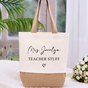 May include: Cream-colored tote bag with a burlap bottom and tan handles. The bag features the text "Mrs. Jocelyn TEACHER STUFF" in black script, with a small heart symbol. A bouquet of white flowers sits to the right.