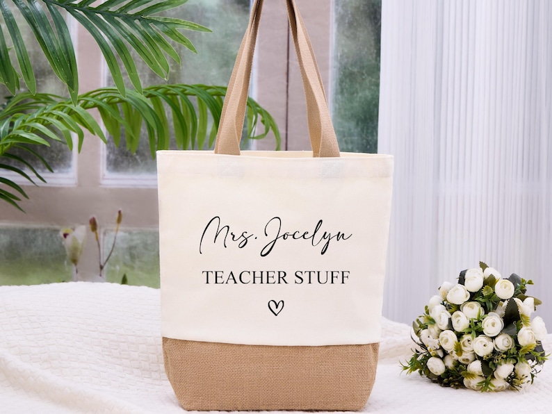 May include: Cream-colored tote bag with a burlap bottom and tan handles. The bag features the text "Mrs. Jocelyn TEACHER STUFF" in black script, with a small heart symbol. A bouquet of white flowers sits to the right.