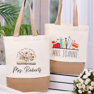 May include: Two canvas tote bags with beige handles and a burlap bottom. One bag features a floral design with the text "Teach them, love them, watch them grow" and "Mrs. Roberts." The other bag has a school supplies graphic and the name "Mrs. Joanne."
