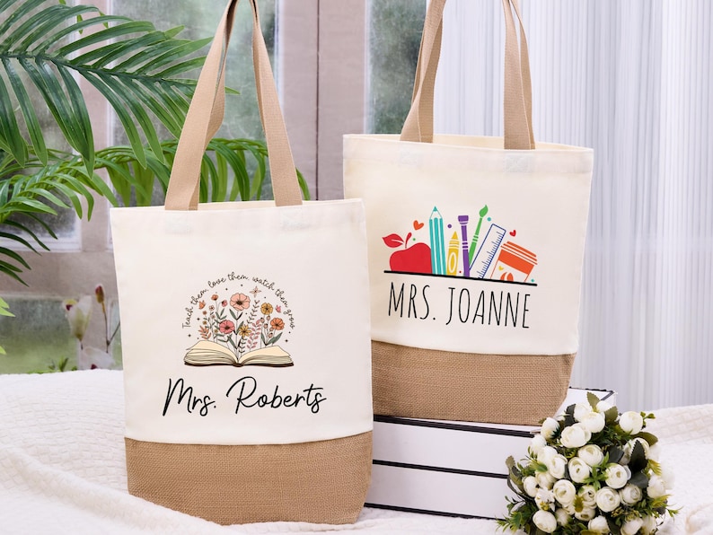 May include: Two canvas tote bags with beige handles and a burlap bottom. One bag features a floral design with the text "Teach them, love them, watch them grow" and "Mrs. Roberts." The other bag has a school supplies graphic and the name "Mrs. Joanne."