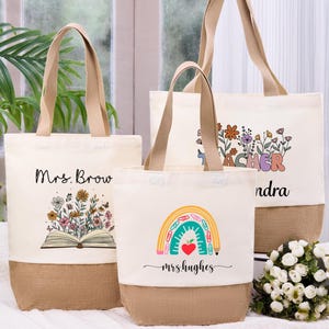 May include: Three canvas tote bags with beige handles and a burlap bottom. One bag features the text "Mrs. Brow" above an open book with flowers. Another says "Teacher" with floral accents. The third says "mrs.hughes" with a rainbow.